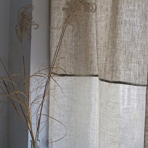 May include: Beige linen curtains with a dark trim hang near a window. A clear glass bottle with dried grasses sits on the windowsill. The curtains have a textured appearance, and the overall aesthetic is minimalist and natural.