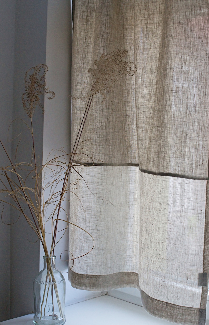 May include: Beige linen curtains with a dark trim hang near a window. A clear glass bottle with dried grasses sits on the windowsill. The curtains have a textured appearance, and the overall aesthetic is minimalist and natural.