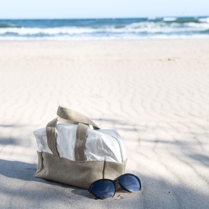 May include: A beige and white canvas tote bag with brown leather handles sits on a sandy beach with a pair of black sunglasses next to it.