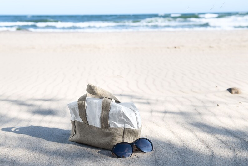 May include: A beige and white canvas tote bag with brown leather handles sits on a sandy beach with a pair of black sunglasses next to it.