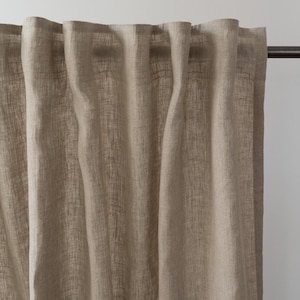 Op de afbeelding: Beige linnen gordijnpaneel dat aan een donkere metalen stang hangt. Het gordijn heeft een textuur met verticale plooien, waardoor het een zachte, vloeiende uitstraling heeft. De neutrale kleur en het eenvoudige ontwerp suggereren een minimalistische of natuurlijke esthetiek.