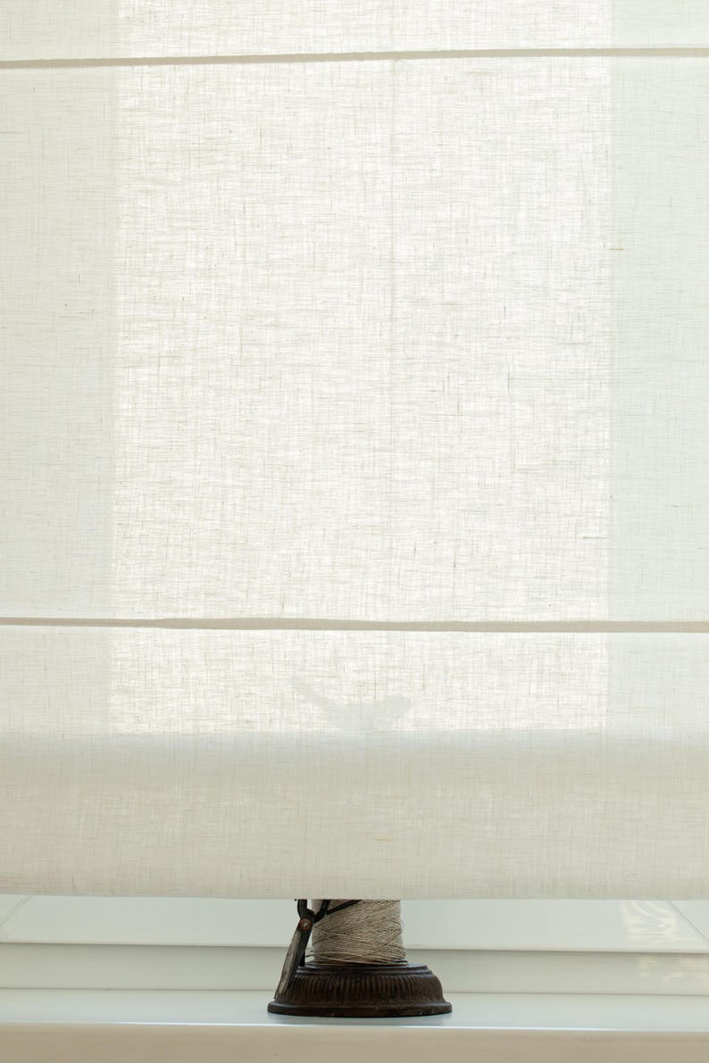 May include: A close-up of a cream-colored Roman shade made of a textured fabric. The shade is partially lowered, revealing a glimpse of the window and a decorative spool at the bottom. The fabric appears to be linen or a similar material.