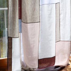 Puede incluir: Una cortina de patchwork con un diseño geométrico. La cortina presenta paneles rectangulares en tonos beige, blanco, rosa y burdeos, delineados con ribetes marrón oscuro. La cortina está hecha de una tela texturizada y cuelga frente a una ventana.