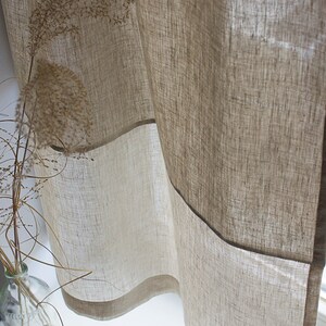 May include: Beige linen curtains with a light brown border, hanging near a window. The curtains are partially transparent, allowing light to filter through. A glass vase with dried plants is visible in the foreground, adding a natural element to the scene.