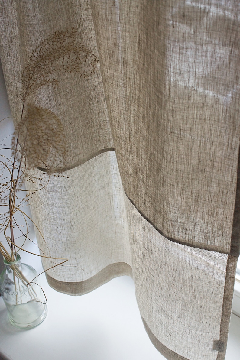 May include: Beige linen curtains with a light brown border, hanging near a window. The curtains are partially transparent, allowing light to filter through. A glass vase with dried plants is visible in the foreground, adding a natural element to the scene.