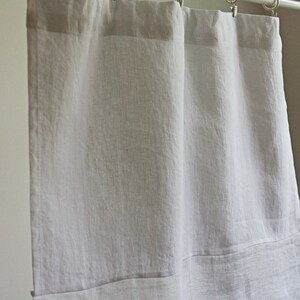 May include: Pale grey linen curtains suspended from a silver curtain rail with metal rings and clips. The curtains have a simple design, with a folded hem at the bottom. The fabric has a slightly textured appearance.