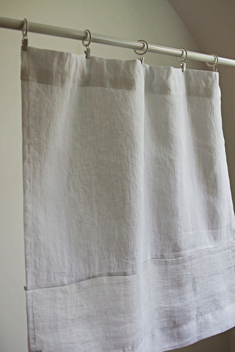 May include: Pale grey linen curtains suspended from a silver curtain rail with metal rings and clips. The curtains have a simple design, with a folded hem at the bottom. The fabric has a slightly textured appearance.