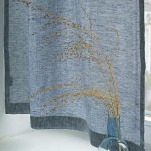 May include: A blue linen curtain with a dark blue border hangs in a window. A vase with dried grass is in front of the curtain.