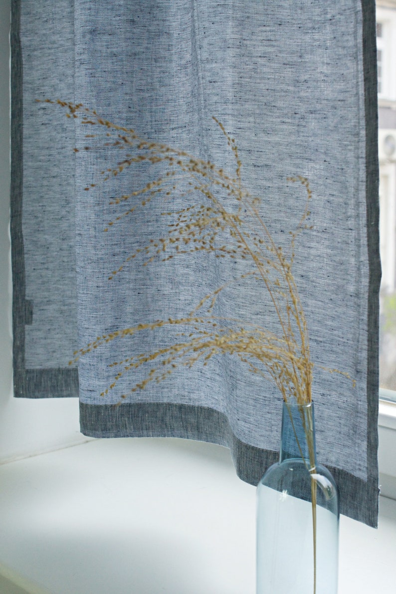 May include: A blue linen curtain with a dark blue border hangs in a window. A vase with dried grass is in front of the curtain.