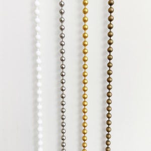 May include: Four beaded chains in white, silver, gold, and bronze colors. Each chain is composed of evenly spaced, round beads. The chains are vertically aligned against a white background, showcasing the variety of metallic and neutral tones.