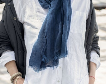 May include: A person wearing a dark blue scarf with frayed edges, draped over a white button-down shirt. The person is also wearing a black jacket, blue jeans, and bracelets. The scarf is the focal point of the image.