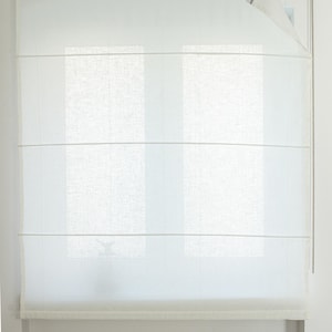 May include: A white Roman shade is mounted on a window. The shade is made of a sheer, light-colored fabric, with horizontal folds. A chain mechanism is visible on the right side. The window behind the shade is partially visible.