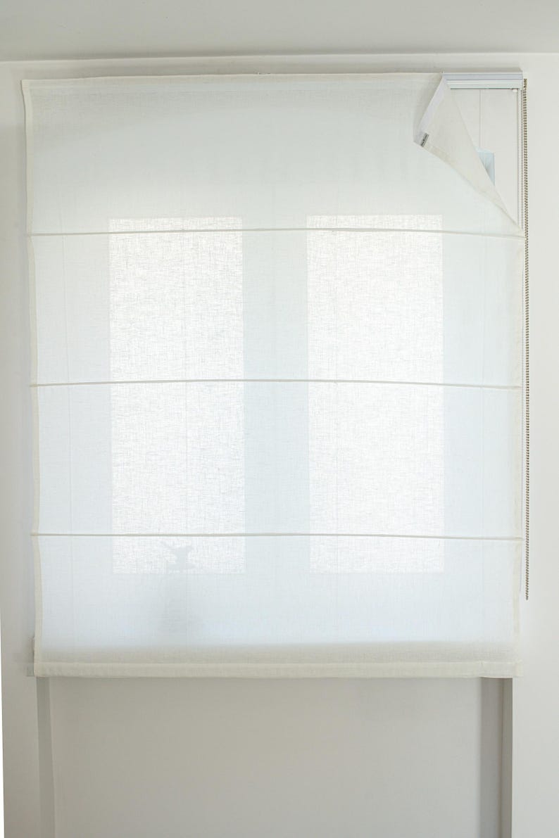 May include: A white Roman shade is mounted on a window. The shade is made of a sheer, light-colored fabric, with horizontal folds. A chain mechanism is visible on the right side. The window behind the shade is partially visible.