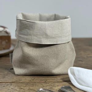 May include: A beige linen bread basket with a folded-over top. The basket sits on a wooden surface, with a white napkin and silverware in the foreground. A loaf of bread is partially visible in the background.