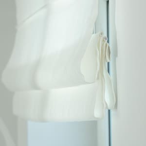 May include: A close-up of a white Roman shade, partially drawn, with soft folds. The shade is made of a light, sheer fabric, and is hanging next to a white wall. The window frame is visible.