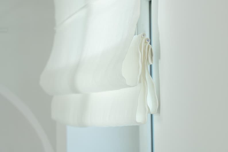 May include: A close-up of a white Roman shade, partially drawn, with soft folds. The shade is made of a light, sheer fabric, and is hanging next to a white wall. The window frame is visible.