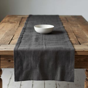 May include: A dark gray linen table runner with a white ceramic bowl on a rustic wooden table. The runner is centered on the table, creating a simple, elegant dining setting. The table has a natural wood grain finish.