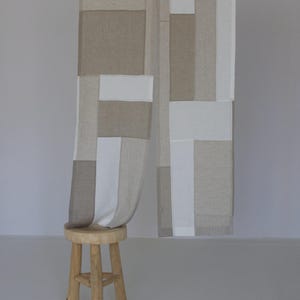 Pode incluir: Uma cortina de patchwork bege e branco com um padrão geométrico pendura em uma haste. A cortina está drapeada sobre um banquinho de madeira.