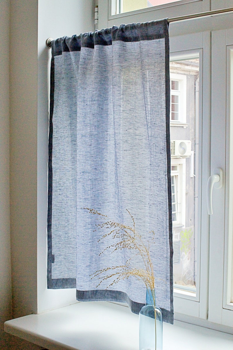 May include: A white linen curtain with a dark gray border hangs in a window. The curtain is sheer and allows light to pass through. A vase with dried flowers sits on the windowsill.