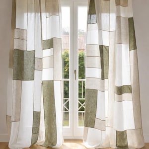 May include: Patchwork-style window curtains in shades of white, beige, and olive green hang in front of a white-framed window. The curtains are made of rectangular fabric panels, creating a geometric pattern. The room has a wooden floor.