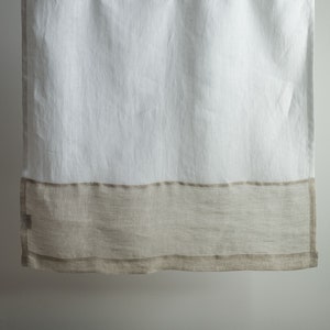 May include: White linen shower curtain with a wide, natural-coloured band at the bottom. The curtain hangs from a white rod with metal hooks. The fabric appears slightly textured, adding to its simple, elegant design. A neutral-toned home decor item.