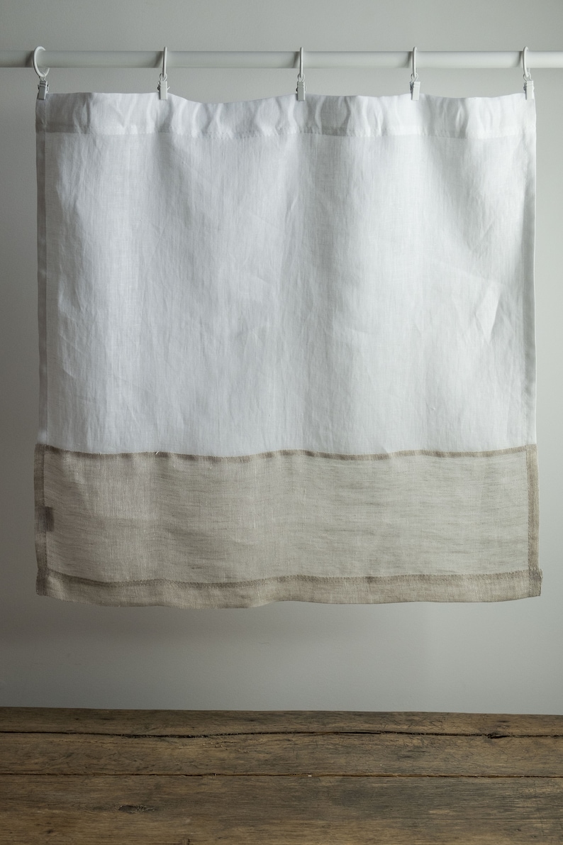 May include: White linen shower curtain with a wide, natural-coloured band at the bottom. The curtain hangs from a white rod with metal hooks. The fabric appears slightly textured, adding to its simple, elegant design. A neutral-toned home decor item.