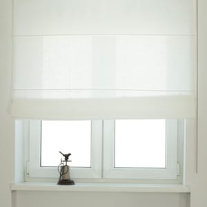 May include: A white Roman shade is installed above a double-pane window. A small decorative bird statue sits on the windowsill. The shade is pulled up, revealing the window. A chain is visible on the right side of the shade.
