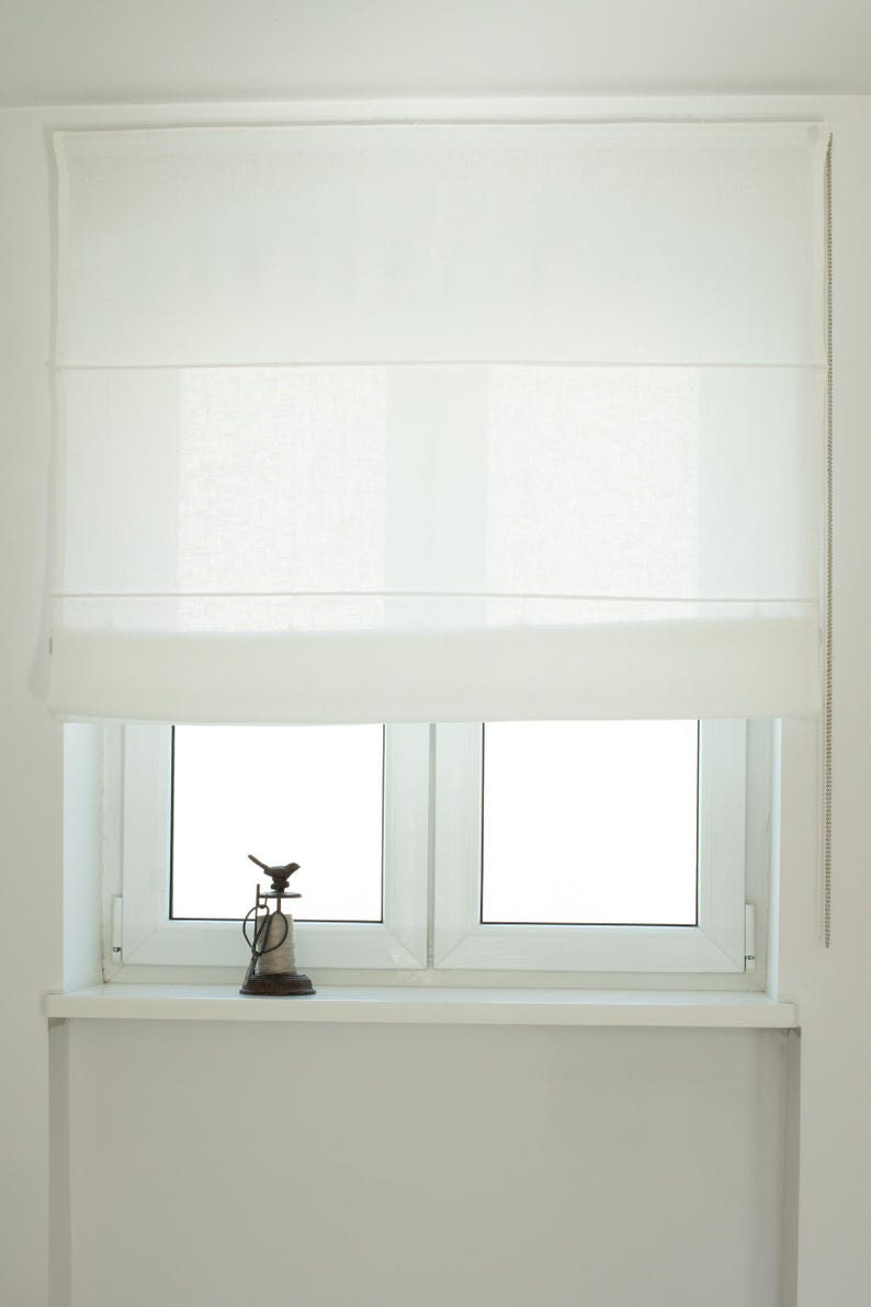 May include: A white Roman shade is installed above a double-pane window. A small decorative bird statue sits on the windowsill. The shade is pulled up, revealing the window. A chain is visible on the right side of the shade.