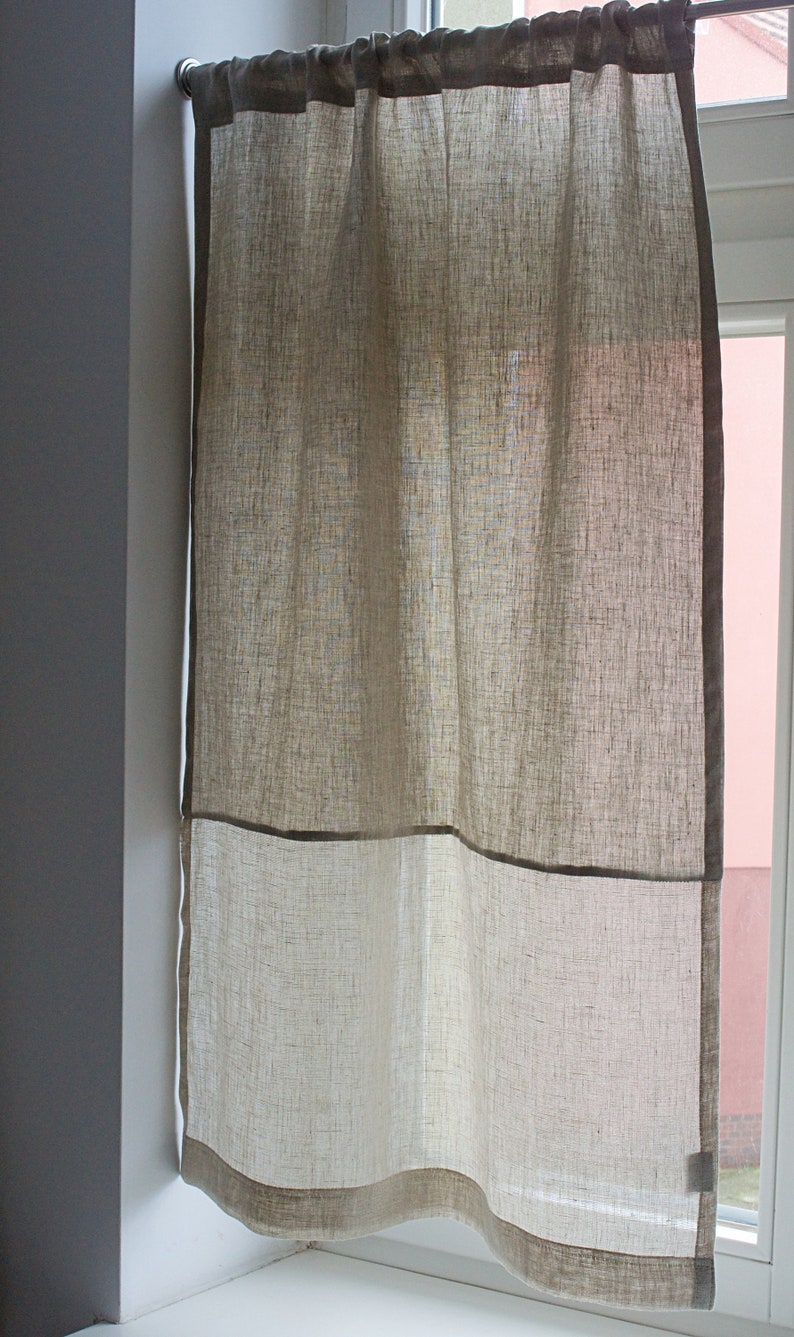 May include: A linen window curtain in shades of beige and off-white. The curtain is divided into two sections, with a darker beige border framing the bottom and top. The fabric has a textured appearance, and it hangs from a rod above a window.