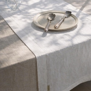 May include: A neutral-toned linen table runner with a matching tablecloth. A silver plate with a spoon and butter knife sits on the runner. A clear glass vase is in the background. The scene is lit by natural light.