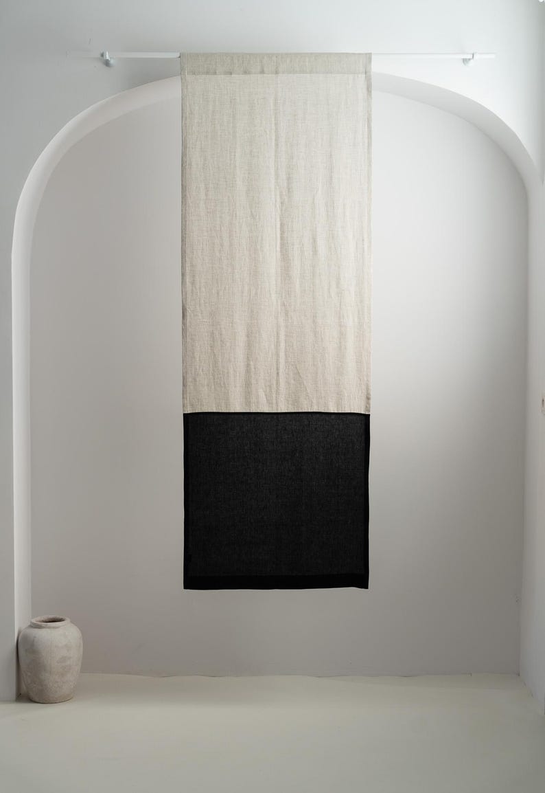 Puede incluir: Una cortina de lino de dos tonos, con una secci&oacute;n superior beige natural y una secci&oacute;n inferior de carb&oacute;n oscuro, cuelga de una barra blanca. La cortina se muestra contra una pared blanca, con una puerta arqueada y un jarr&oacute;n decorativo en la esquina.