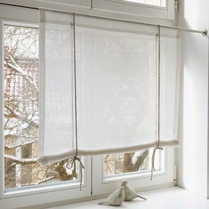 May include: A light-coloured linen window blind hangs in front of a window. The blind is held up by a metal rod and has a rolled bottom. Dark brown cords are used to raise and lower the blind. Two white ceramic bird figurines sit on the windowsill.