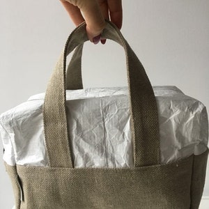 May include: A small, beige canvas tote bag with a white paper lining and two thick canvas handles.