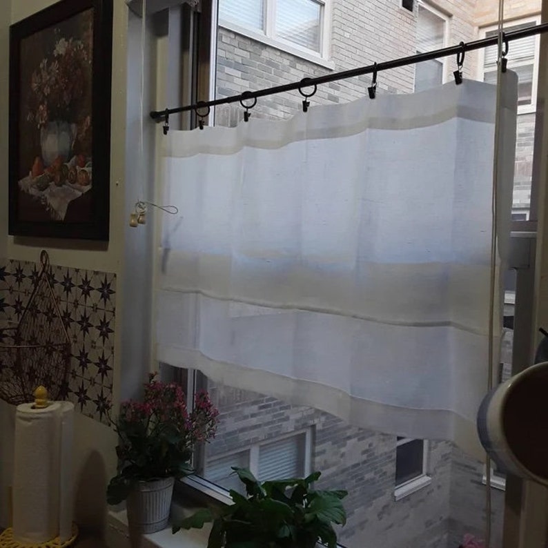 May include: A white, semi-transparent curtain is suspended from a black rod with rings in a window. The curtain features a layered design with a lighter border. A framed painting and potted plants are visible in the background, with a view of a brick building.