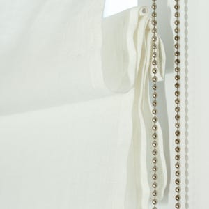 May include: Close-up of a white Roman shade with a textured fabric. A beaded chain hangs on the right side. The shade is partially drawn, revealing its layered construction and soft folds. The overall aesthetic is minimalist and elegant.