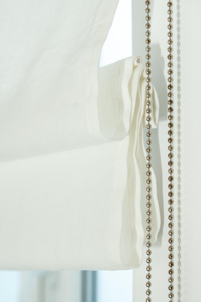 May include: Close-up of a white Roman shade with a textured fabric. A beaded chain hangs on the right side. The shade is partially drawn, revealing its layered construction and soft folds. The overall aesthetic is minimalist and elegant.