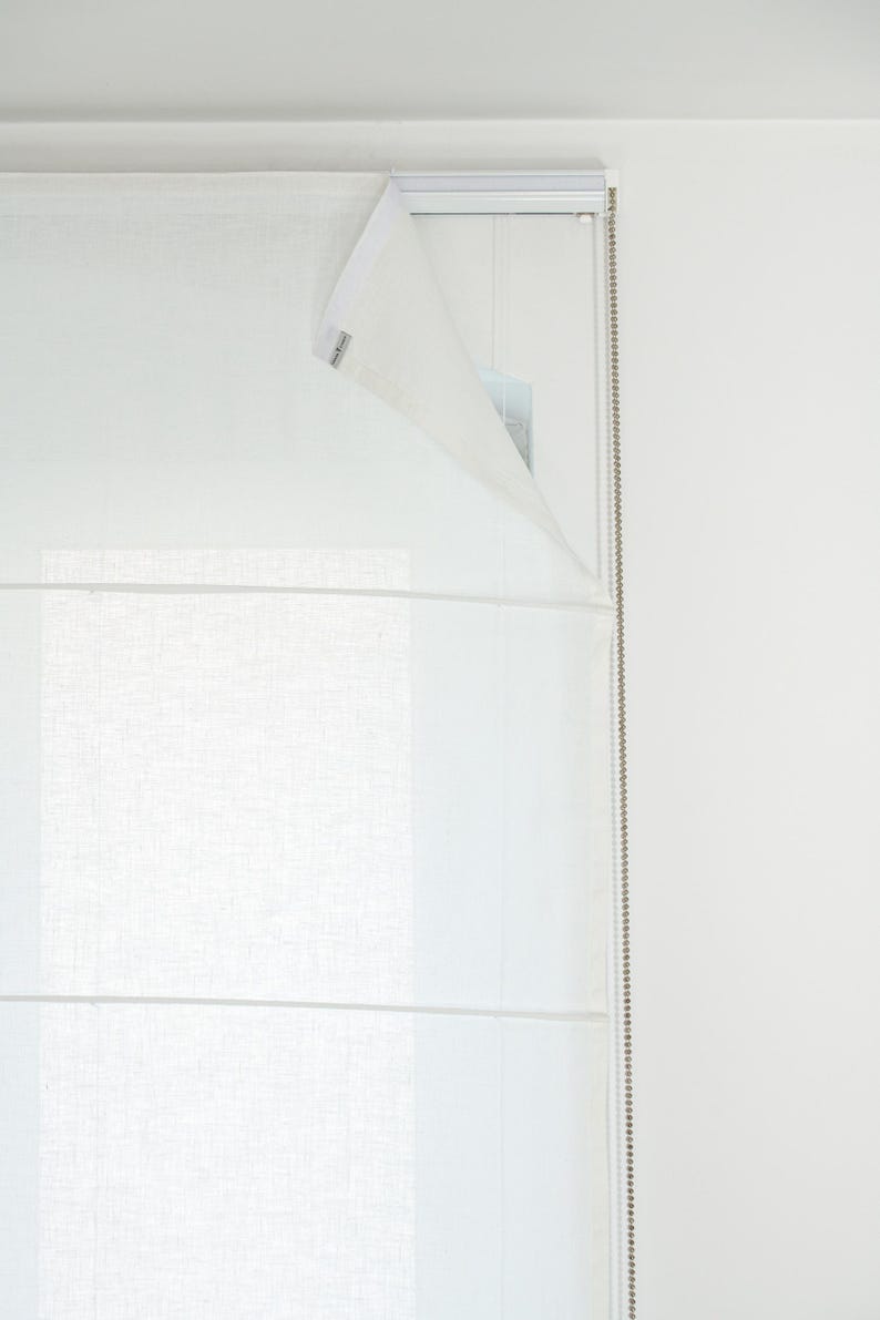 May include: A white Roman shade is partially open, revealing a glimpse of the window and the mechanism. The shade has horizontal folds and a textured appearance. A beaded chain hangs on the right side.