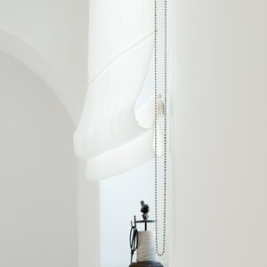 May include: A bright, minimalist interior scene featuring a white Roman shade, a beaded chain, and a vintage spool holder. The spool holder is made of dark metal and holds a spool of thread. The scene is bathed in natural light.