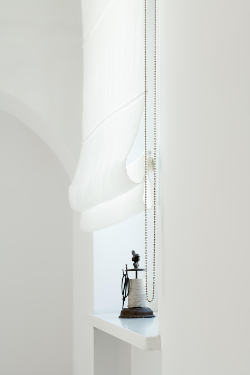 May include: A bright, minimalist interior scene featuring a white Roman shade, a beaded chain, and a vintage spool holder. The spool holder is made of dark metal and holds a spool of thread. The scene is bathed in natural light.