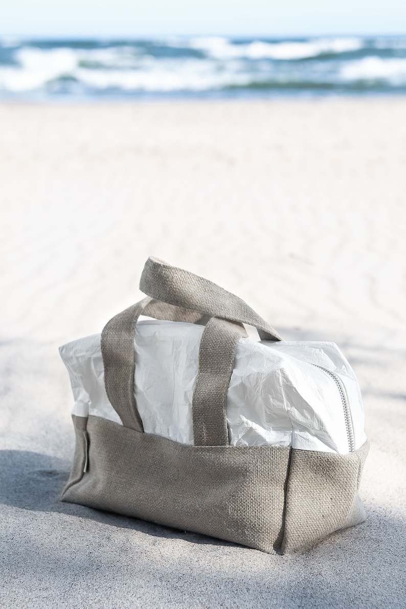May include: A white canvas tote bag with brown canvas straps. The bag is sitting on a sandy beach.
