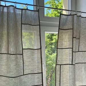 May include: Neutral-toned linen curtains with a geometric design. The curtains are divided into rectangular panels with dark brown borders. The curtains hang from a silver rod in front of a window with a view of trees and a blue sky.