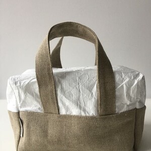 May include: A beige linen tote bag with a white paper lining. The bag has two handles and a rectangular shape.