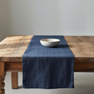 Op de afbeelding: Een marineblauwe linnen tafelloper ligt op een rustieke houten tafel. Een witte keramische kom staat op de loper. De tafel heeft een natuurlijke houten afwerking.