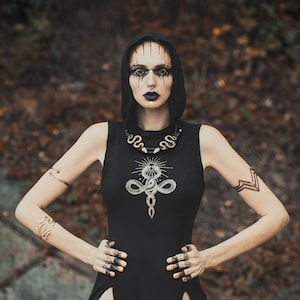 Op de afbeelding: Een vrouw in een zwarte jurk met capuchon met een slangendesign en gouden sieraden. Ze heeft donkere make-up en een serieuze uitdrukking.