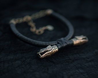 May include: A black leather choker necklace with two gold snake head clasps. The necklace is on a black textured surface.