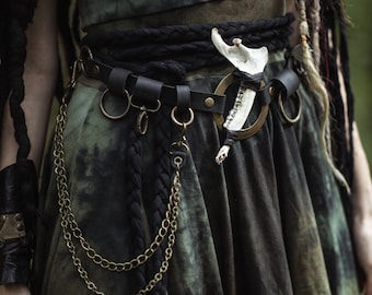 May include: A black leather belt with a large silver buckle and a bone charm. The belt is worn over a green and black tie-dye skirt with a black braided rope.