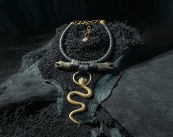 May include: A black leather choker necklace with a gold-colored snake pendant. The necklace features gold-colored chain and decorative snake head accents. The jewelry is displayed on a dark, textured surface.