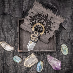 May include: A clear quartz crystal point wrapped in a black snake design with a gold ring and a wooden box with a mandala design.