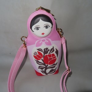 May include: Pink crossbody bag shaped like a Russian nesting doll, featuring a painted face and floral design. The bag has a gold zipper and strap attachments, with a detachable pink strap.