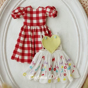 May include: Two doll dresses. One is red and white gingham with puffed sleeves. The other is white with a yellow heart and colorful embroidery. The text "Craftsman Doll" is visible in the image.